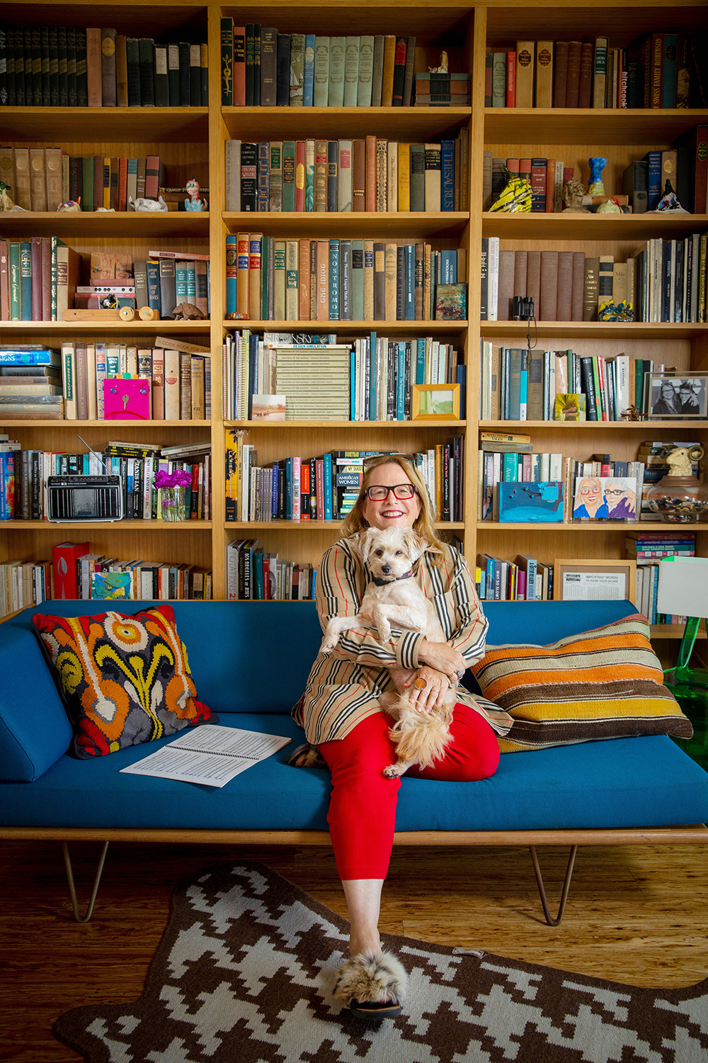 Laura Karpman sits on a bench holding a dog in front of a tall wall of bookshelves.